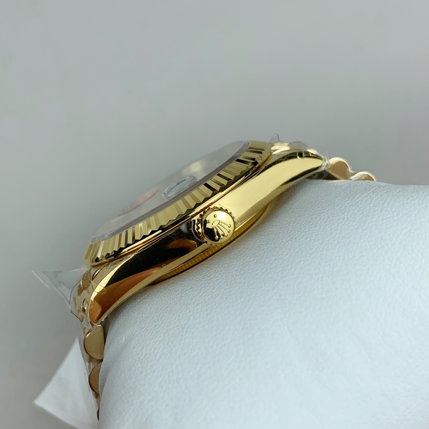Angled side view of gold-toned wristwatch resting on white textured surface showing polished gold case, fluted bezel, textured crown with Rolex logo, domed crystal, and Oyster bracelet links