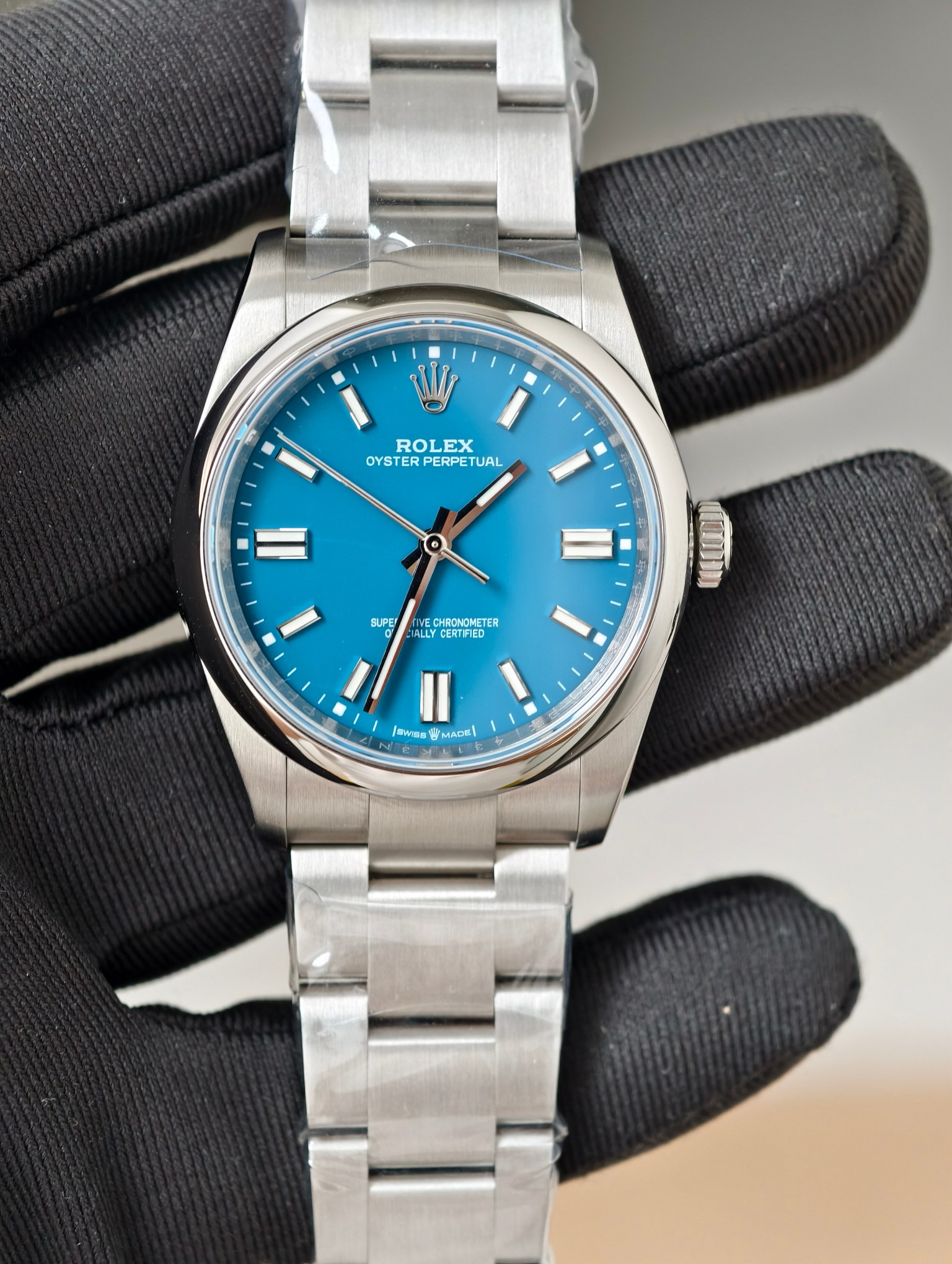 Close-up of Rolex Oyster Perpetual with vibrant blue sunburst dial held in black-gloved hand, showing polished stainless steel case, Oyster bracelet, and detailed markers