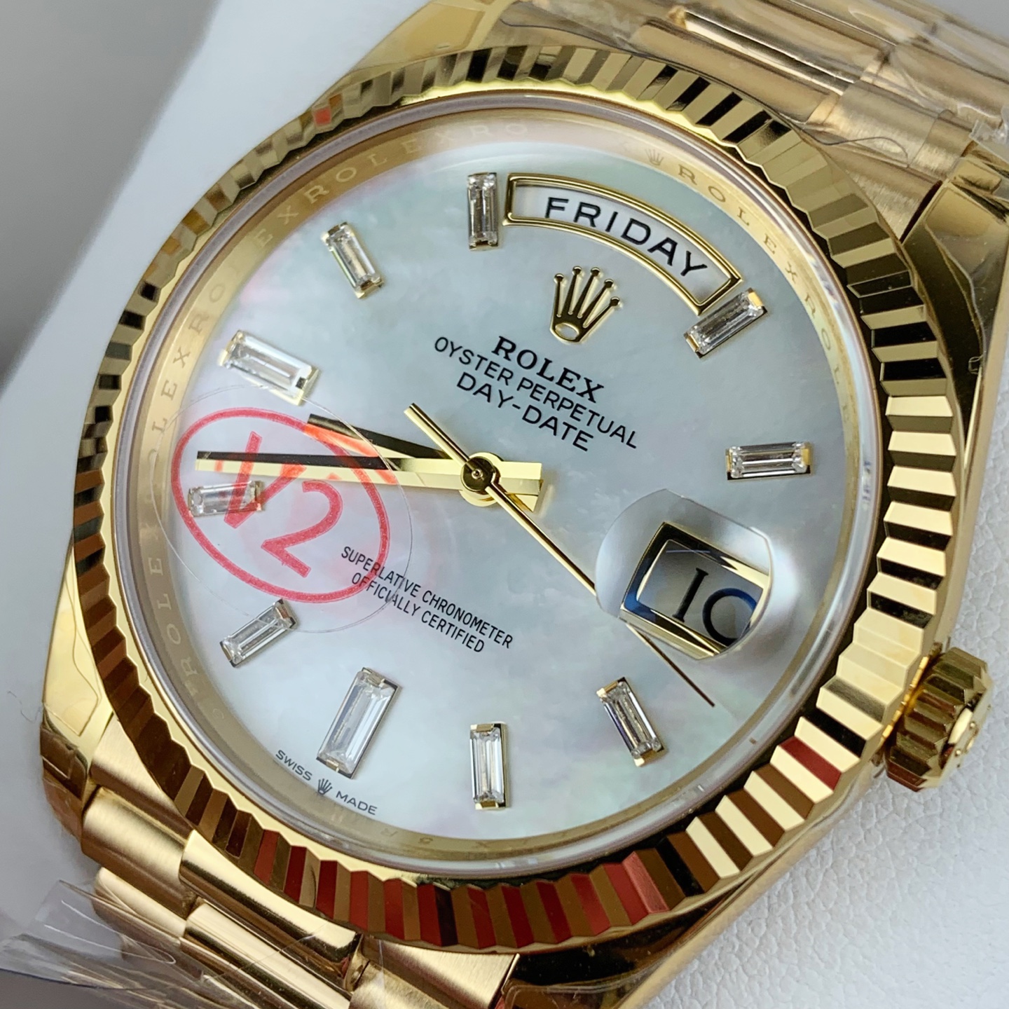 Close-up photograph of gold Rolex Oyster Perpetual Day-Date wristwatch with mother-of-pearl dial, baguette-cut diamond hour markers, day window showing FRIDAY, date window with Cyclops lens showing 10, and red quality stamp