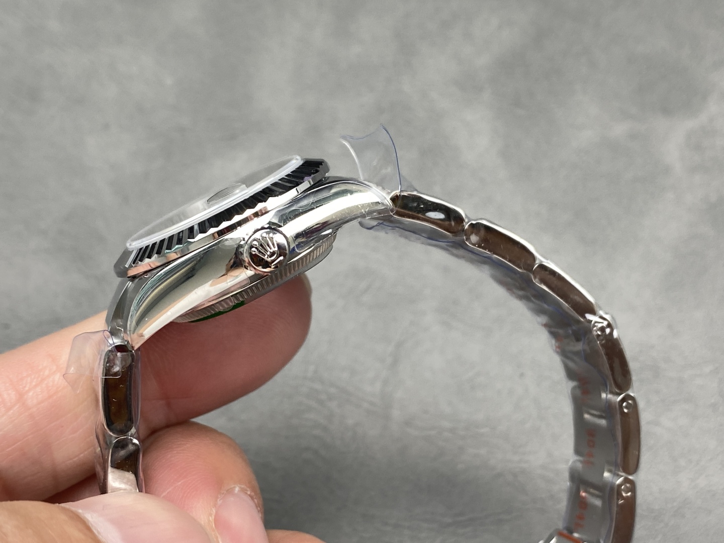Side view of the stainless steel Rolex Datejust replica showing the fluted screw-down crown with Rolex logo engraving, polished case lugs, and Oyster bracelet with protective plastic film