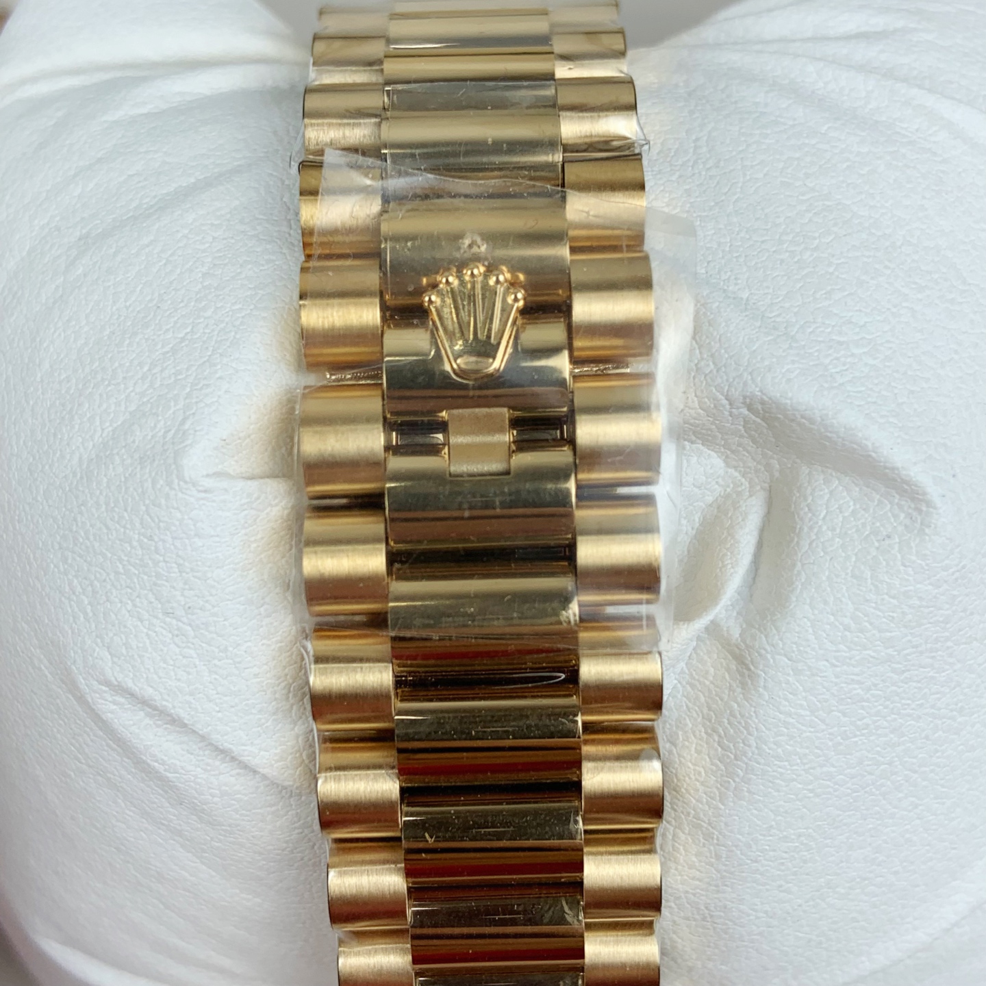 Back of gold Oyster bracelet resting on white textured surface showing polished and brushed links, central clasp with engraved Rolex crown logo, and partial protective plastic film