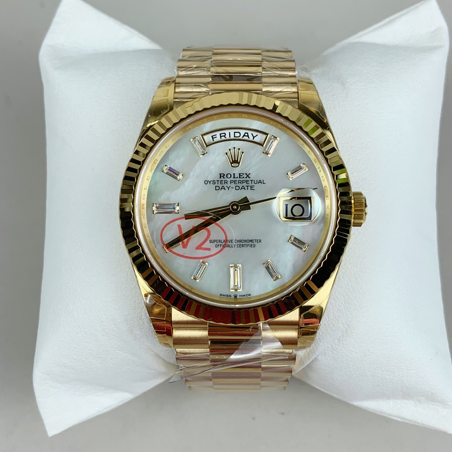 Gold-toned Rolex Day-Date wristwatch presented on a white padded display stand featuring mother-of-pearl dial with day and date windows, baguette-cut diamond hour markers, fluted bezel, and red V2 stamp