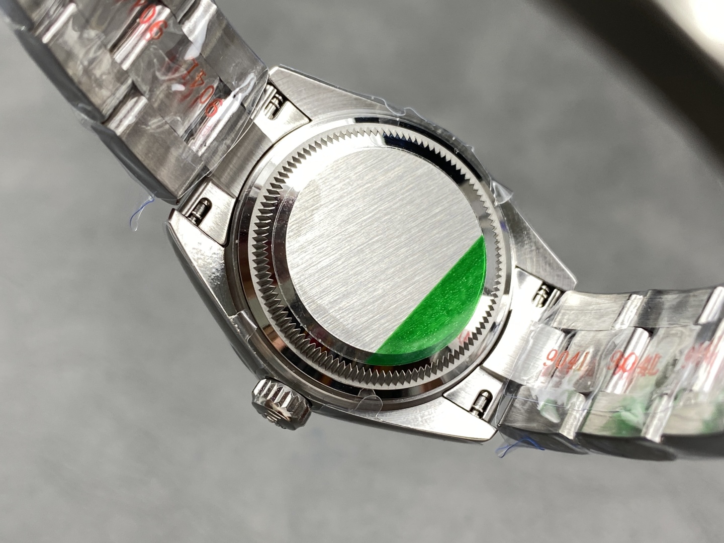 Rear view of the Rolex replica caseback showing brushed stainless steel finish with radial pattern, green protective sticker, and bracelet links with red markings and plastic wrap