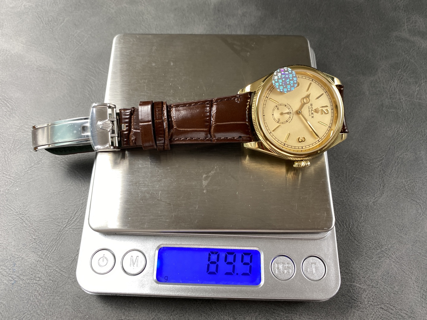 Replica Rolex watch on digital scale showing 89.9g weight, gold dial with blue gem bezel and brown leather strap