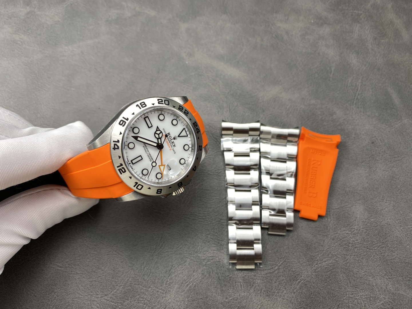 Full view of Rolex Explorer II replica with orange rubber strap held by gloved hand, alongside stainless steel bracelet and strap changing tool