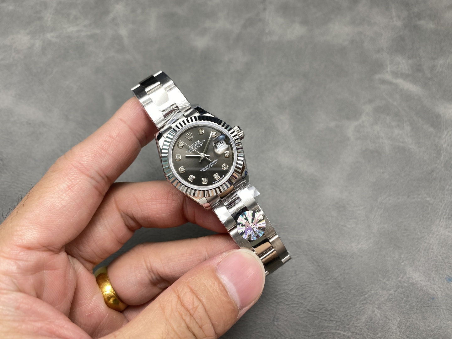 High-end Rolex Datejust replica featuring a dark gray sunburst dial with twelve diamond hour markers, fluted bezel, and polished Oyster bracelet, held in a hand against a textured background