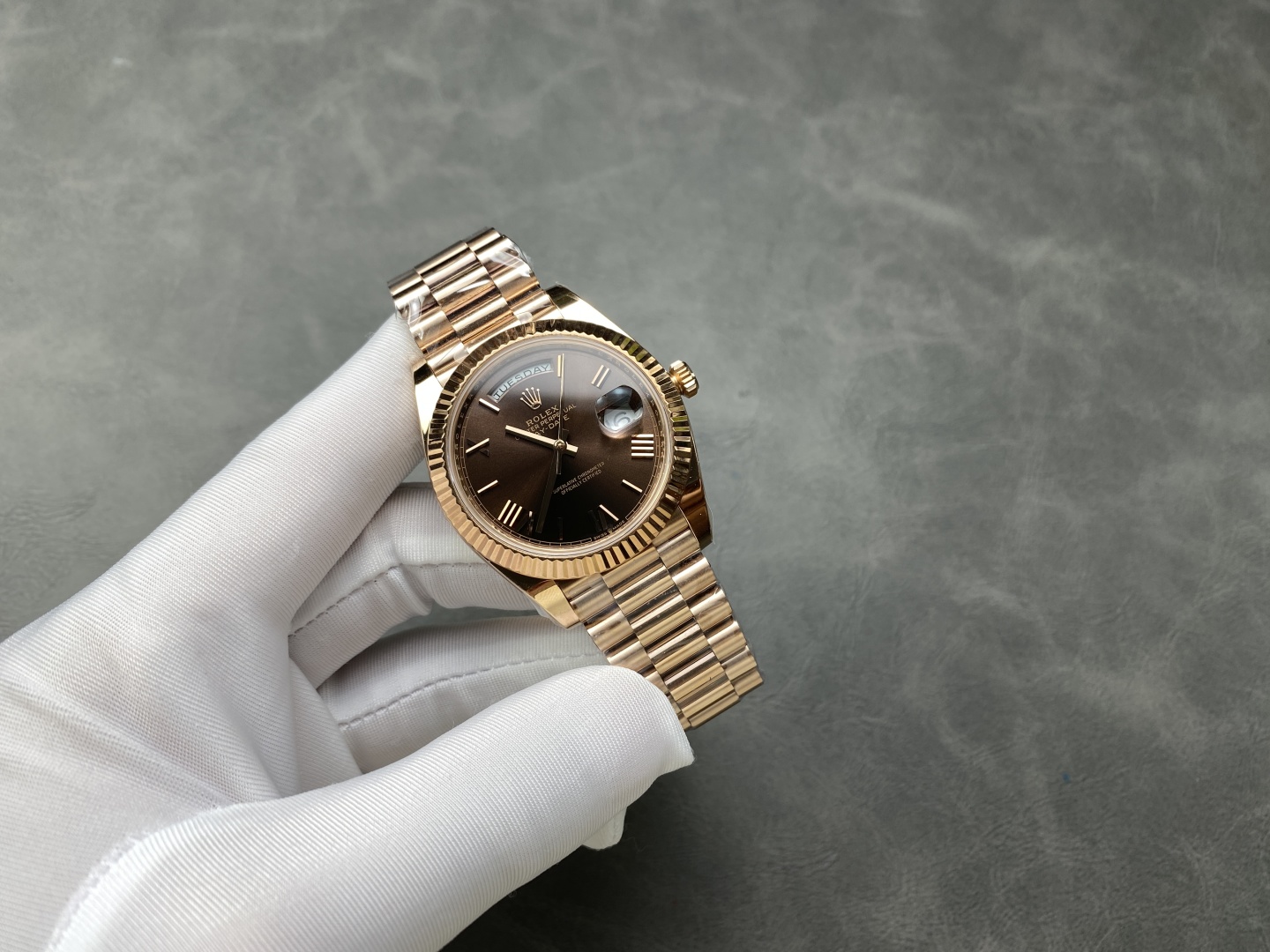 Rose gold Rolex Day-Date replica watch held with white glove on grey surface
