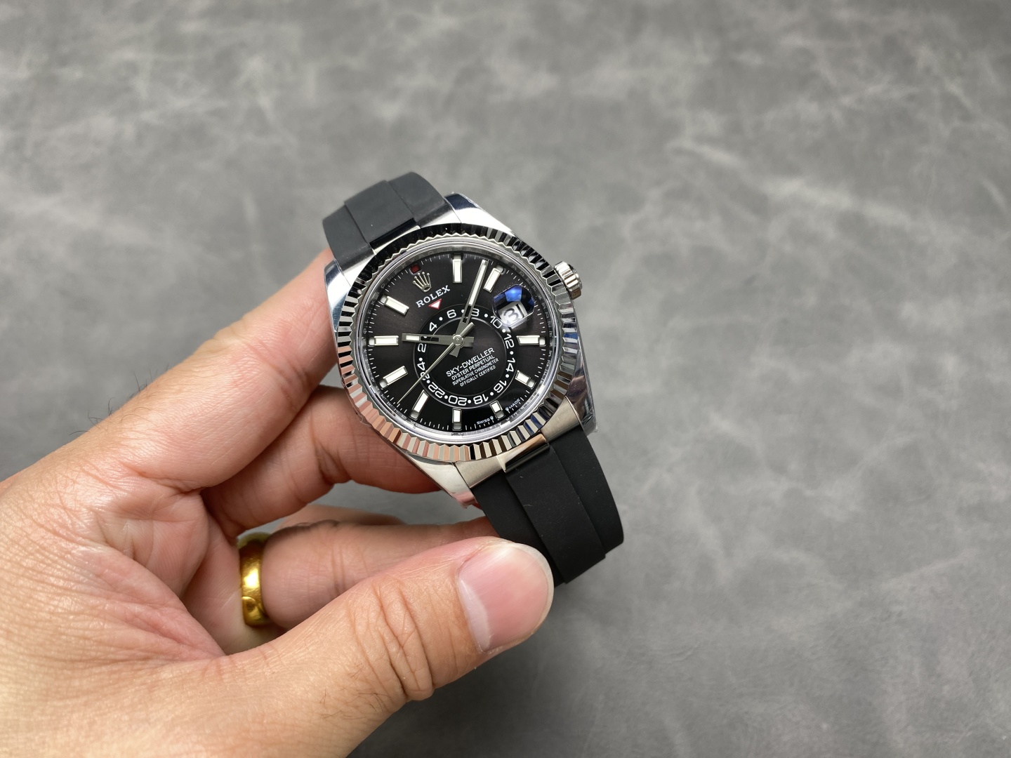 Hand holding black dial Rolex Sky-Dweller replica with rubber strap on gray surface